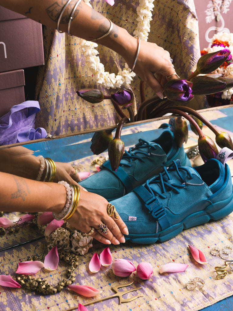 The Kali Blue sneakers are displayed in an altar setting, held by the model's hands. The space around the sneakers include pink lotus petals and the stems of unbloomed blue waterlilies.