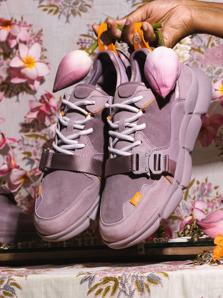 The Purple Sunset sneakers are displayed in an altar setting, held by the model's hands, against a floral fabric background.