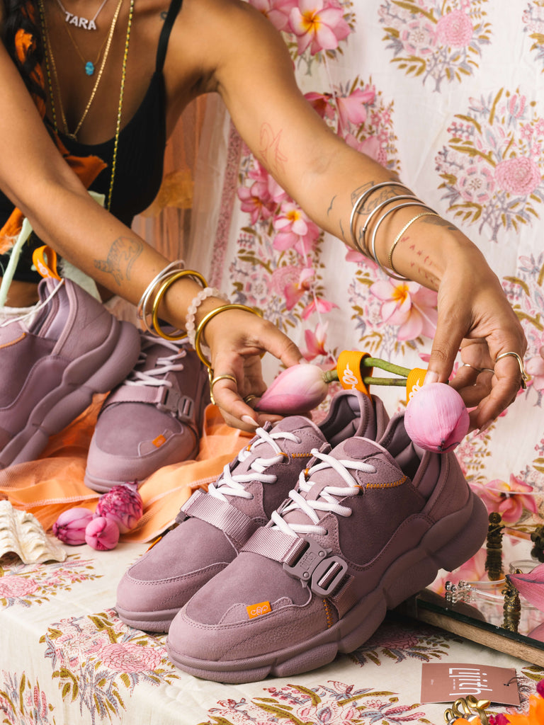 The Purple Sunset sneakers are displayed in an altar setting, held by the model's hands, against a floral fabric background.