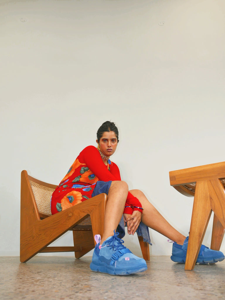 Model Elina Banerjee is seated leaning forward in a Le Corbusier-style chair, wearing her Kali Blue sneakers. Location is Chandigarh.