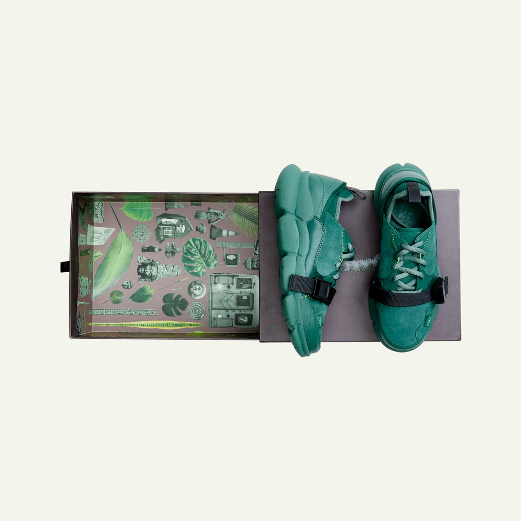 A pair of Caudal Lure sneakers in the color Concrete Jungle Green, placed on top of a chocolate brown colored Lilith NYC shoebox. The sole uses a Vibram tooling called Plump in the Pantone color Duck Green. The upper materials are made up of various shades of green in the materials suede, pebbled leather and neoprene. The Lilith NYC wordmark logo is printed at the back of the sneaker.