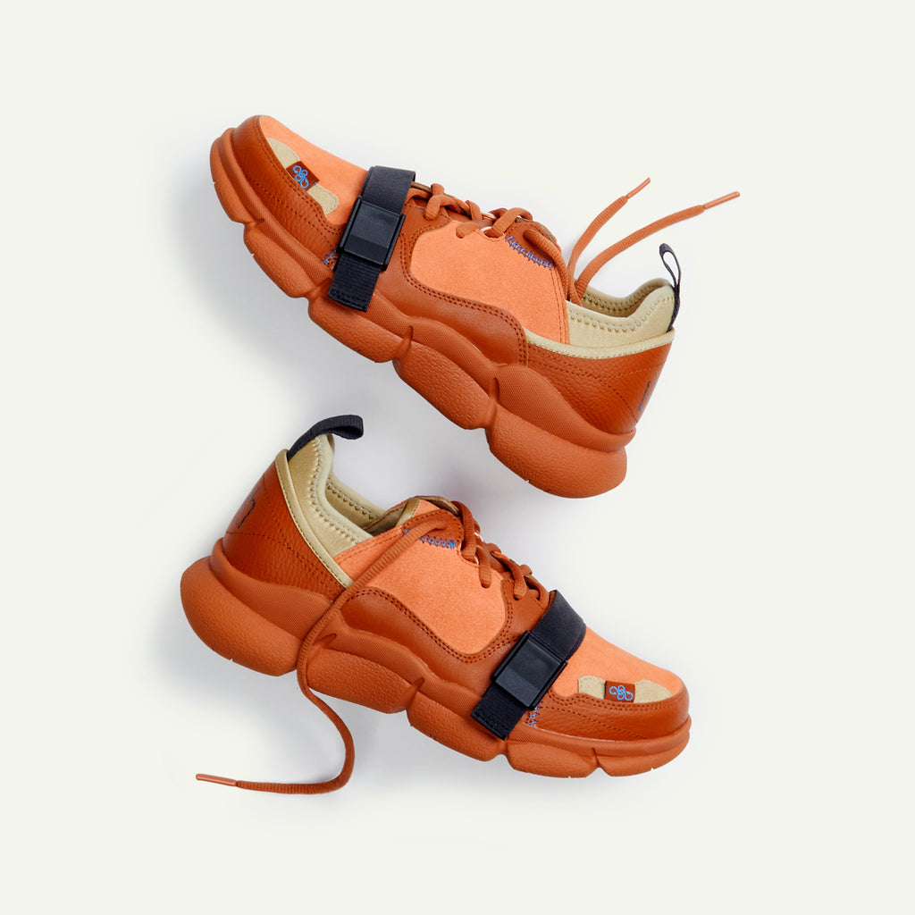 A pair of Caudal Lure sneakers in the color Amberlou Brick. The sole uses a Vibram tooling called Plump in the Pantone color Umber. The upper materials are made up of various shades of orange in the materials suede, pebbled leather and neoprene. The Lilith NYC wordmark logo is printed at the back of the sneaker.